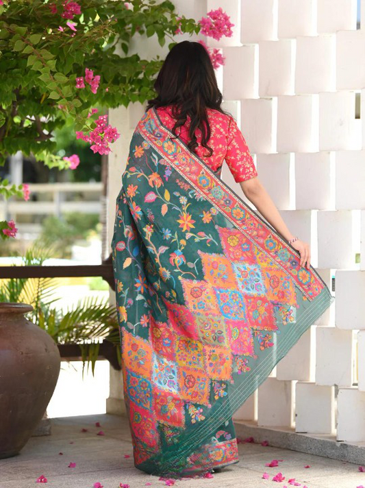 Woman wearing a colorful saree with a floral pattern, standing outdoors near a white wall and greenery.