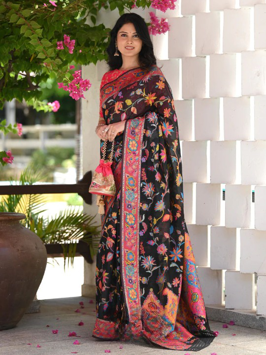 Woman wearing a colorful saree with a floral pattern, standing against a white brick wall with pink flowers.