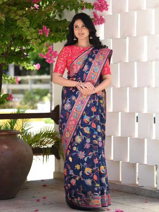Woman wearing a blue and pink saree with a floral pattern, standing in front of a white brick wall.