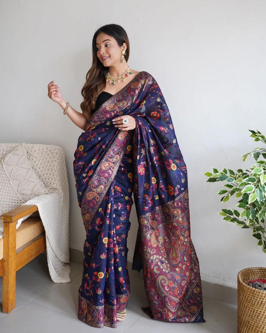 Woman wearing a dark blue saree with floral patterns in a room with a chair and plant.