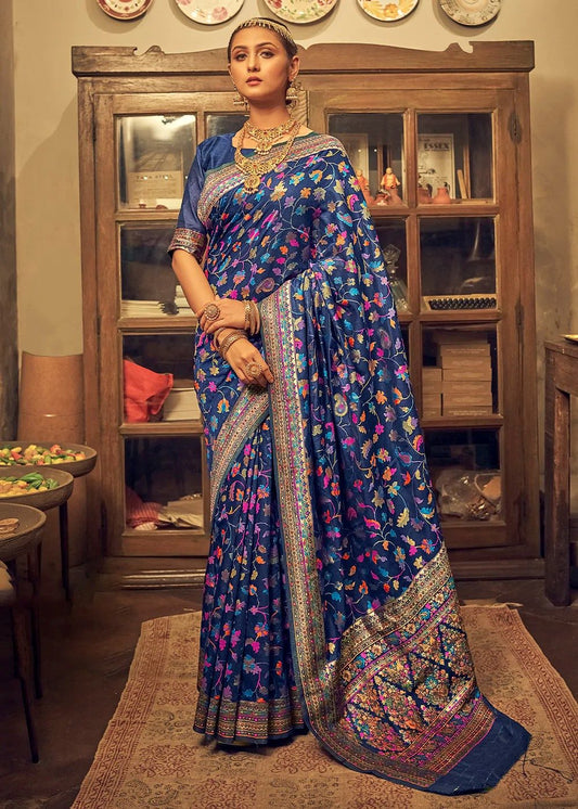 Woman in a blue saree with floral patterns standing in a room with wooden furniture.