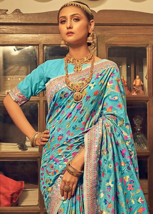 Woman in a blue saree with floral patterns and gold jewelry in an indoor setting.