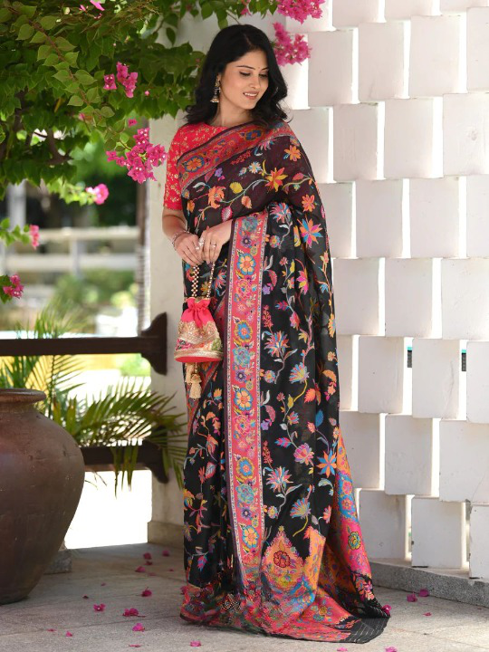 Woman wearing a black floral saree with a white wall and greenery in the background